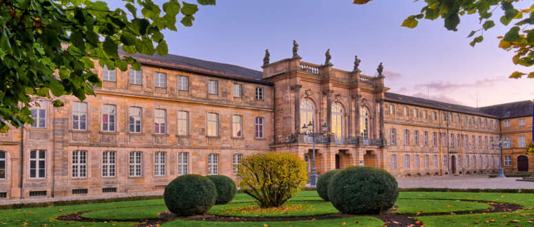 Hinter einem gepflegten Rasen und kreisförmigen Hecken erhebt sich ein historisches Steingebäude mit Statuen auf dem Dach, eingerahmt von grünen Bäumen mit Herbstlaub unter einem pastellfarbenen Himmel.