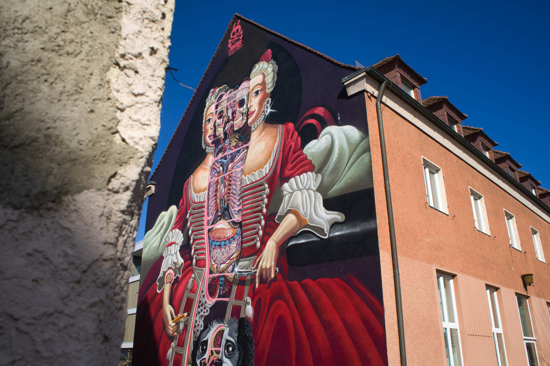 Ein Wandgemälde an einem Gebäude zeigt eine Frau in einem roten Kleid, deren Organe auf stilisierte, anatomische Weise sichtbar sind, vor einem dunklen Hintergrund unter einem strahlend blauen Himmel.