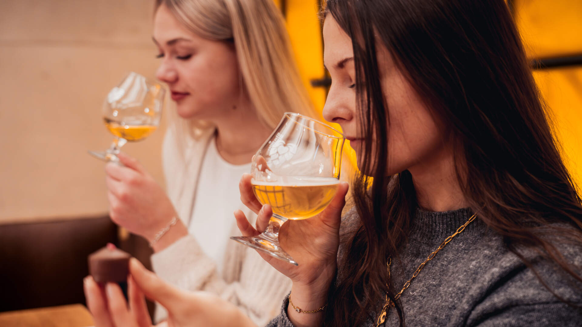 Zwei Frauen sitzen nebeneinander, jede hält ein Glas Bier in der Hand und genießt dessen Duft. Eine der Frauen hält zudem ein Stück Schokolade in der Hand. Beide scheinen in einer warm beleuchteten Umgebung eine Verkostung zu genießen.
