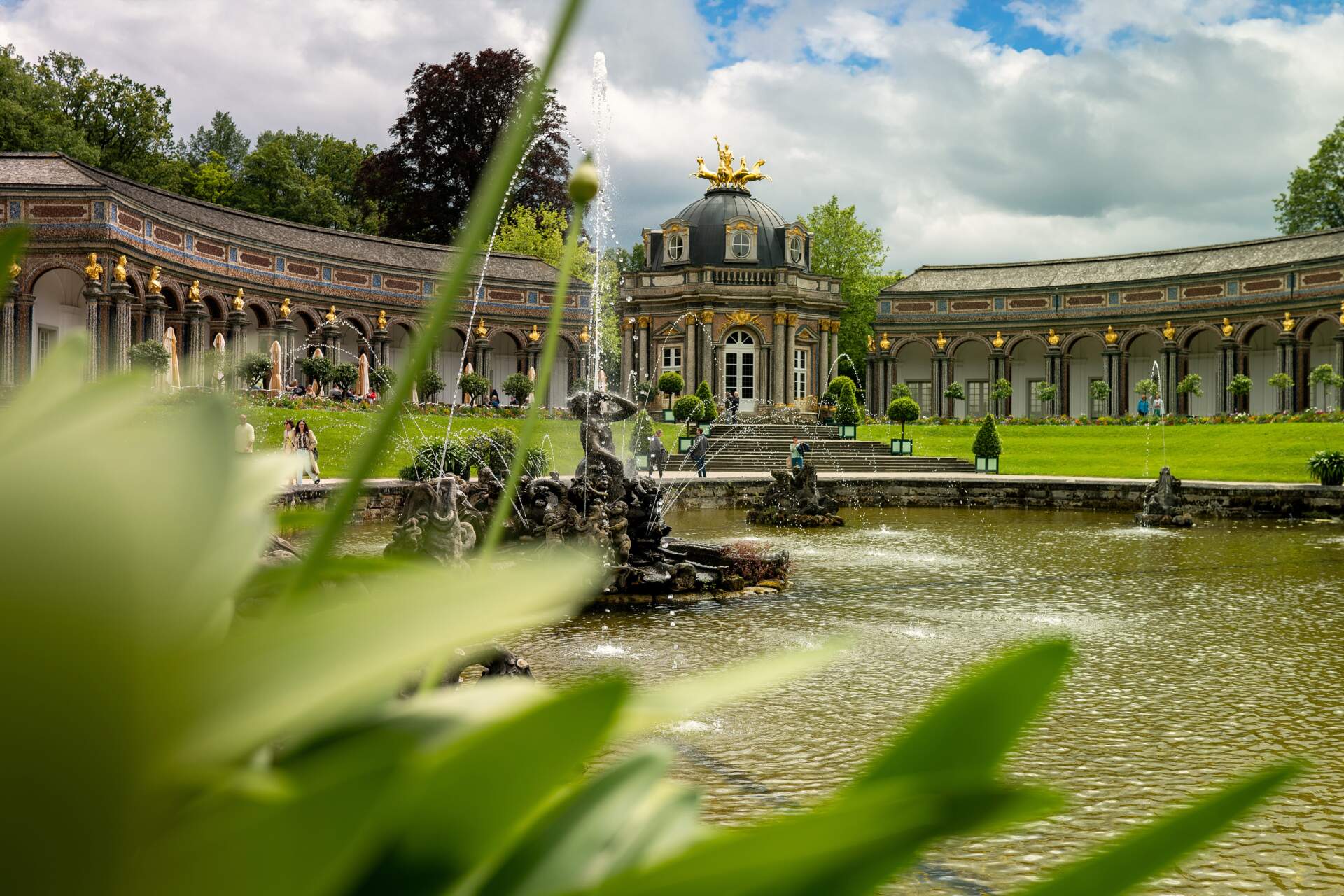 Barockpalast mit kunstvollen goldenen Statuen, geschwungener Kolonnade und einem Springbrunnen in einem Teich, gesehen durch grünes Laub im Vordergrund an einem teilweise bewölkten Tag.