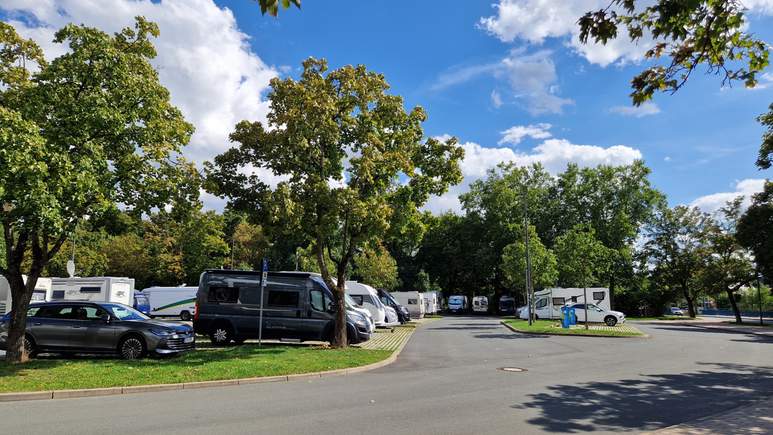 Ein Parkplatz mit mehreren Wohnmobilen und Autos, die unter grünen Bäumen vor einem teilweise bewölkten blauen Himmel stehen, bietet großzügige Wohnmobilstellplätze, auf denen Reisende einen ruhigen Zwischenstopp in der Natur genießen können.