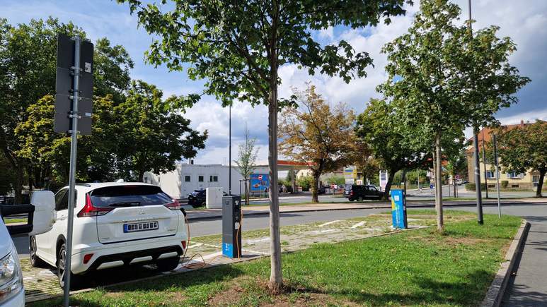 Zwei Elektroautos stehen an Ladestationen in einer von Bäumen gesäumten Straße und spiegeln das Engagement des Viertels für Nachhaltigkeit wider. Der Himmel ist teilweise bewölkt, und im Hintergrund sind Gebäude und geparkte Autos zu sehen. Grünes Gras und Bäume säumen den Gehweg und schaffen eine angenehme Atmosphäre, wie man sie oft in der Nähe moderner Wohnmobilstellplätze vorfindet.