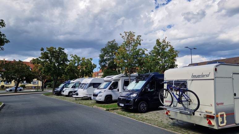 Mehrere Wohnmobile und Wohnwagen stehen in einer Reihe am Straßenrand und nutzen die praktischen Wohnmobilstellplätze. An einem der Fahrzeuge sind hinten zwei Fahrräder befestigt. Bäume säumen den Gehweg, und Wolken bedecken den Himmel.