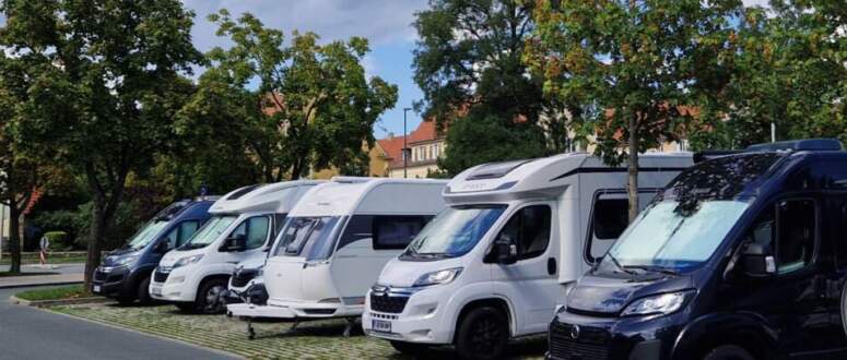 Mehrere Wohnmobile und Wohnwagen stehen in einer Reihe am Straßenrand und nutzen die praktischen Wohnmobilstellplätze. An einem der Fahrzeuge sind hinten zwei Fahrräder befestigt. Bäume säumen den Gehweg, und Wolken bedecken den Himmel.