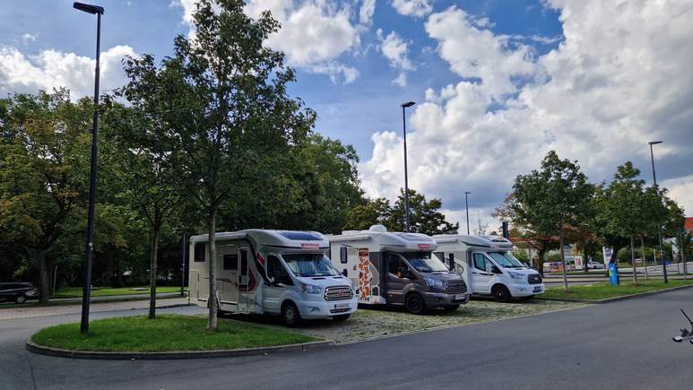 Drei Wohnmobile stehen nebeneinander auf ausgewiesenen Wohnmobilstellplätzen auf einem Parkplatz neben einer Straße, umgeben von Bäumen, Gras und Straßenlaternen. Der Himmel darüber ist teilweise bewölkt, und die Szene wirkt ruhig.