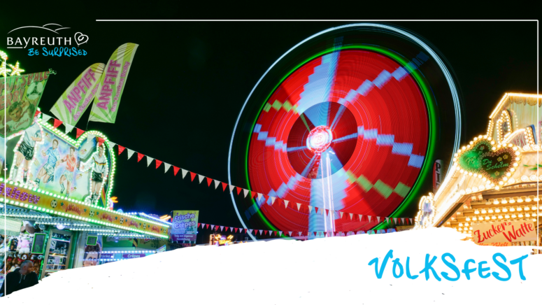 Ein Riesenrad, das nachts auf einem Jahrmarkt mit roten und grünen Lichtern beleuchtet ist; dazu sind Spielstände, Fahnen und der Schriftzug Volksfest zu sehen.