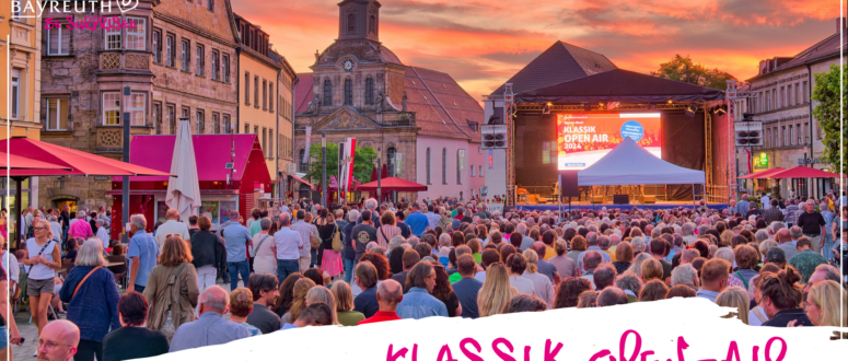 Auf einem Stadtplatz versammelt sich eine große Menschenmenge zu einem Klassik Open Air-Konzert bei Sonnenuntergang, das an die Bayreuther Festspiele erinnert.