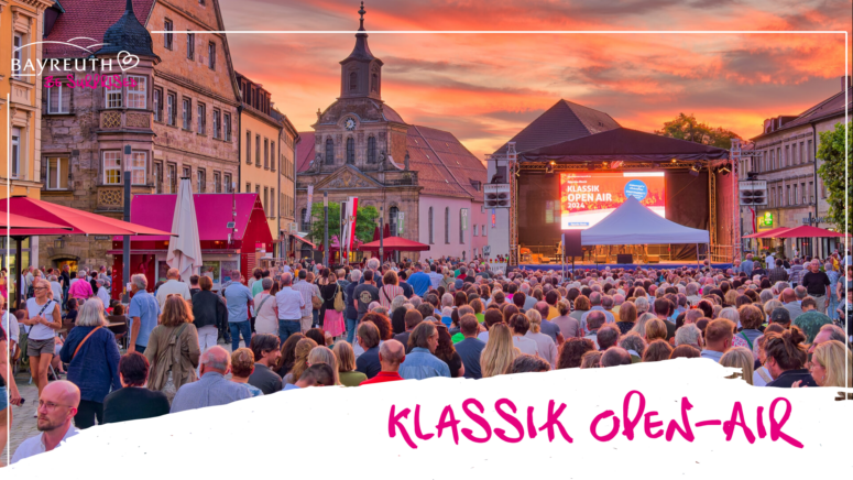 Auf einem Stadtplatz versammelt sich eine große Menschenmenge zu einem Klassik Open Air-Konzert bei Sonnenuntergang, das an die Bayreuther Festspiele erinnert.