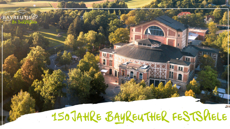 Luftaufnahme des von Bäumen umgebenen Bayreuther Festspielhauses, unten mit dem Schriftzug 150 Jahre Bayreuther Festspiele.