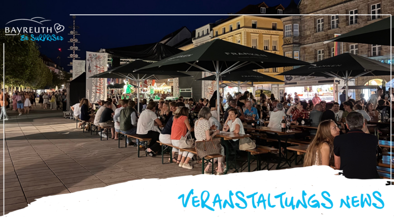 Bei einem nächtlichen Straßenfest im Rahmen der Bayreuther Festspiele sitzen die Menschen an langen Tischen unter dunklen Sonnenschirmen, während im Hintergrund eine Band auf einer Bühne spielt.
