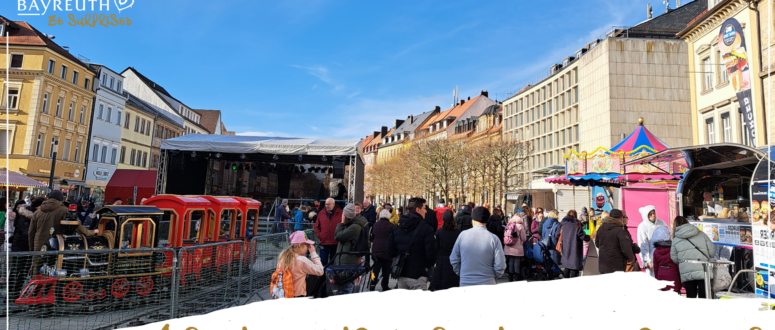 Auf einem Stadtplatz versammelt sich eine Menschenmenge zu einem Faschingsumzug, bei dem ein kleiner roter Kinderzug, eine Bühne und ein Karussell zu sehen sind; am unteren Bildrand ist der Schriftzug Vereine, Künstler und Schausteller eingeblendet.