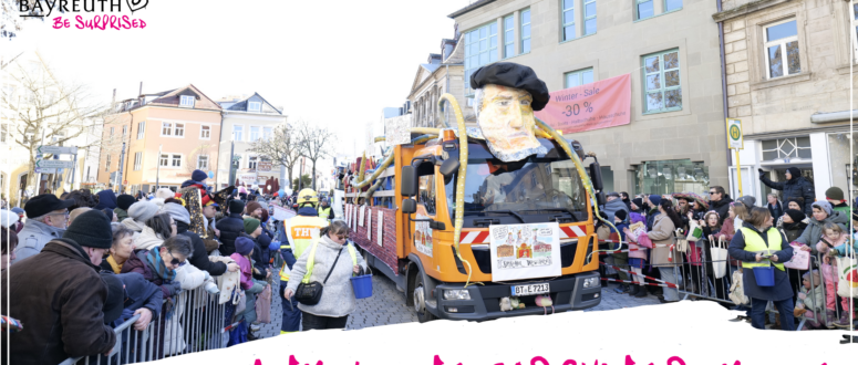 Ein orangefarbener Lastwagen mit einem großen Pappmaché-Kopf, der eine schwarze Baskenmütze trägt, fährt durch die Menge eines Faschingsumzugs in einer deutschen Stadt.