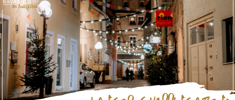 Eine schmale Kopfsteinpflasterstraße bei Nacht, geschmückt mit Lichterketten, mit Weihnachtsbäumen und dem Ladenschild von Rion, auf dem die Aufschriften Bayreuth – Lass dich überraschen und Langer Shoppingabend zu lesen sind.