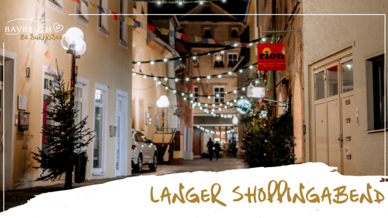 Eine schmale Kopfsteinpflasterstraße bei Nacht, geschmückt mit Lichterketten, mit Weihnachtsbäumen und dem Ladenschild von Rion, auf dem die Aufschriften Bayreuth – Lass dich überraschen und Langer Shoppingabend zu lesen sind.
