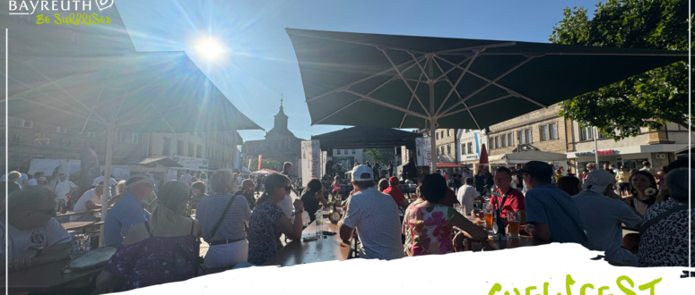 Bei einem Weinfest im Freien sitzen die Menschen an langen Tischen unter großen Sonnenschirmen, im Hintergrund sind eine Bühne und ein Kirchturm zu sehen.