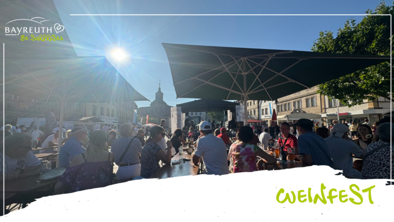 Bei einem Weinfest im Freien sitzen die Menschen an langen Tischen unter großen Sonnenschirmen, im Hintergrund sind eine Bühne und ein Kirchturm zu sehen.