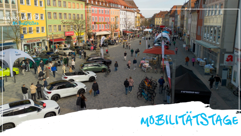 Aufnahme aus der Vogelperspektive eines Stadtplatzes während der Mobilitätstage, mit geparkten Autos, Fahrrädern, Markenzelten und Menschen, die zwischen bunten Gebäuden umhergehen.