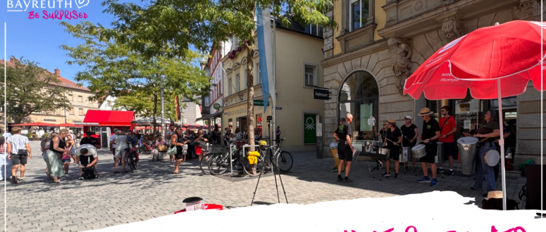 Eine Band, die auf einem mit Kopfsteinpflaster gepflasterten Stadtplatz mit Passanten und Ladenfronten Schlagzeug spielt, unter dem Logo Bayreuth Be Surprised und dem Text Weitere Events.