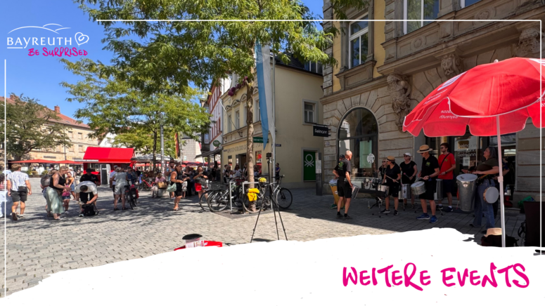 Eine Band, die auf einem mit Kopfsteinpflaster gepflasterten Stadtplatz mit Passanten und Ladenfronten Schlagzeug spielt, unter dem Logo Bayreuth Be Surprised und dem Text Weitere Events.