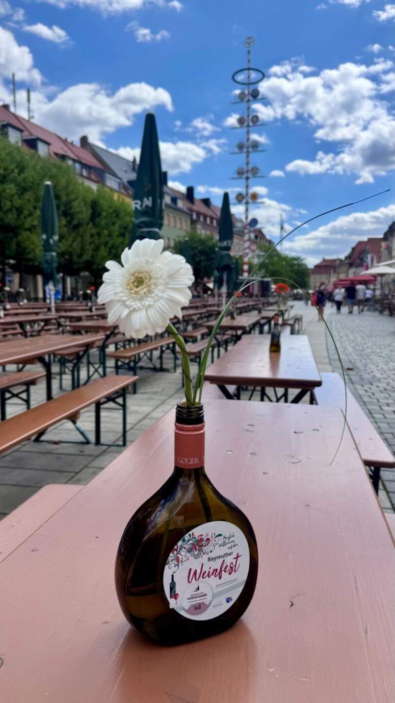 Eine weiße Gerbera in einer Weinflaschenvase auf einem hölzernen Picknicktisch bei einem Weinfest im Freien.