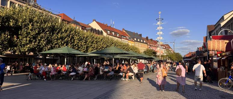 Menschen, die während eines Weinfestes auf einem sonnigen Stadtplatz, der von Gebäuden und einem Maibaum gesäumt ist, an Tischen unter großen grünen Sonnenschirmen sitzen.