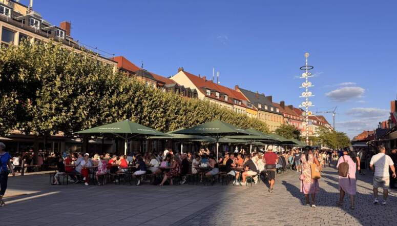 Menschen, die während eines Weinfestes auf einem sonnigen Stadtplatz, der von Gebäuden und einem Maibaum gesäumt ist, an Tischen unter großen grünen Sonnenschirmen sitzen.