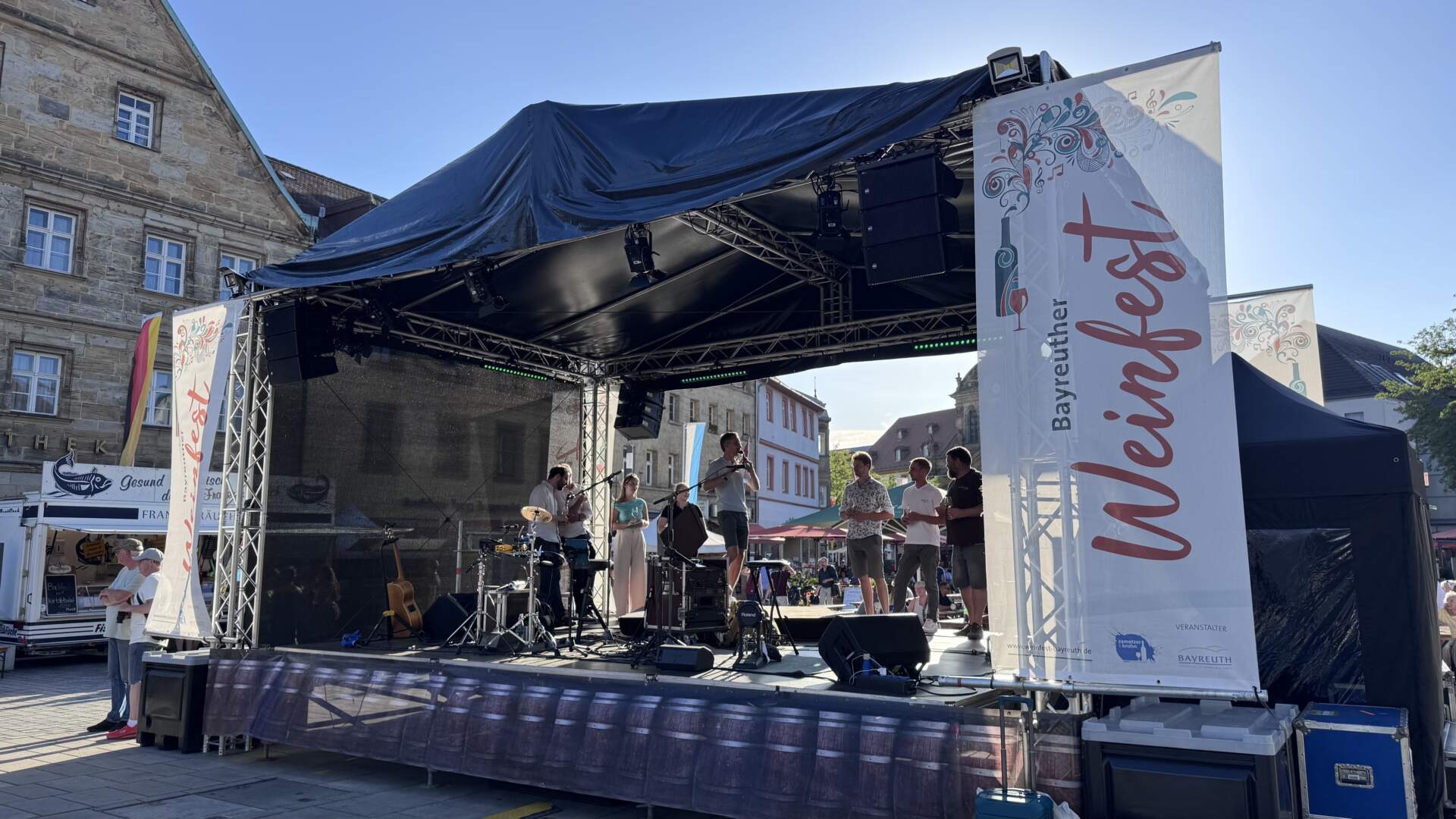 Auf einer Bühne tritt während des Bayreuther Weinfests eine Band auf; an der vorderen Ecke hängt ein großes Werbebanner.