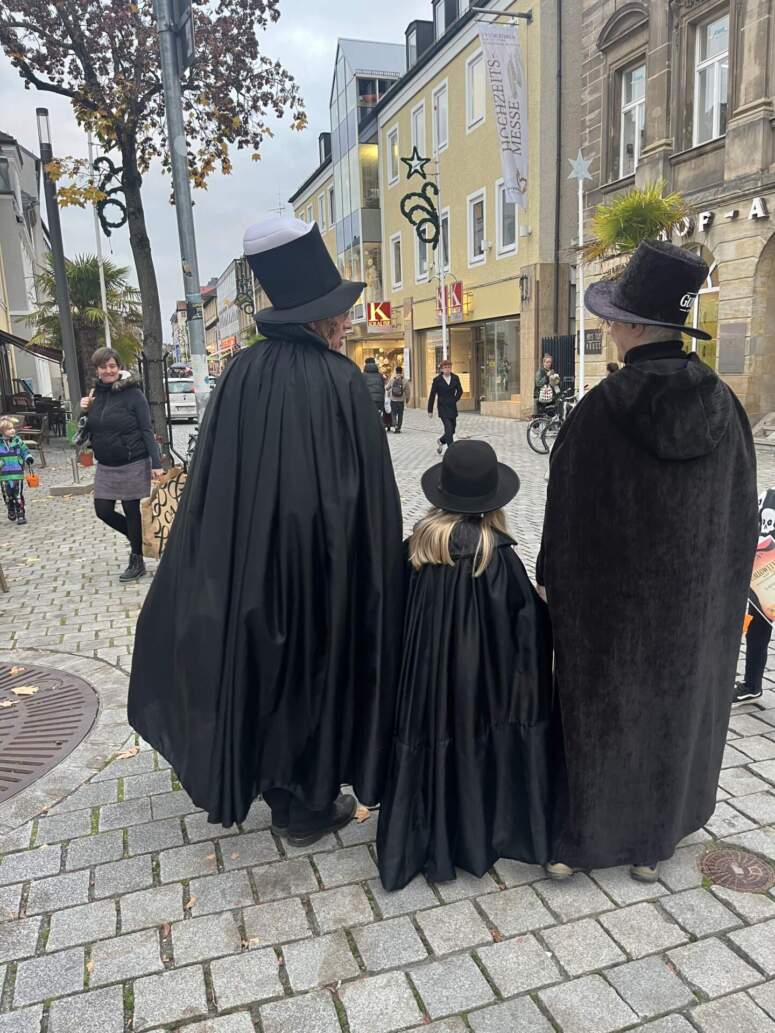 Zwei Erwachsene und ein Kind, die alle schwarze Umhänge und Zylinder tragen, gehen eine mit Kopfsteinpflaster gepflasterte Fußgängerzone entlang.