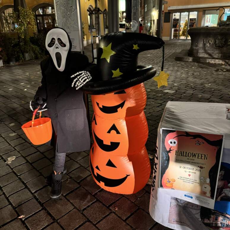 Ein Kind mit einer Ghostface-Maske und einem schwarzen Mantel steht neben einer aufblasbaren Kürbislaterne und einem Schild, das auf eine Halloween-Veranstaltung in Bayreuth hinweist.