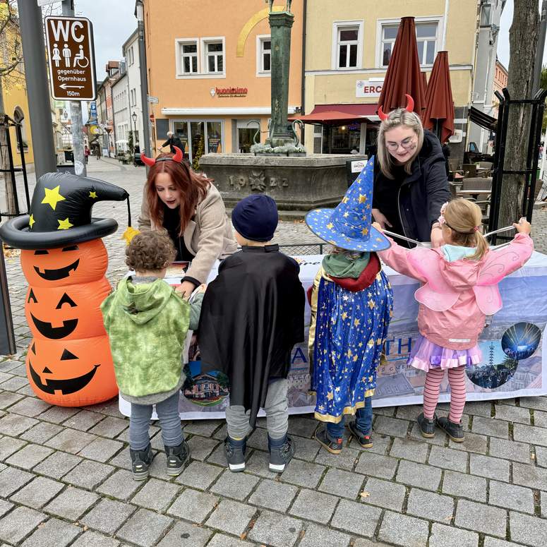 Zwei Frauen mit Teufelshörnern verteilen Süßigkeiten an vier Kinder in Halloween-Kostümen, die an einem Tisch auf einem Stadtplatz stehen.