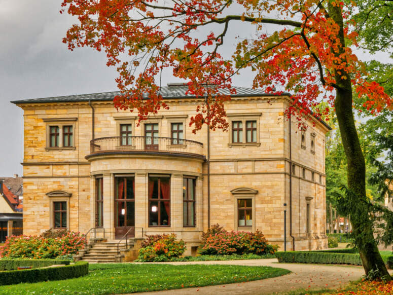 Ein zweistöckiges Herrenhaus aus Stein mit einem geschwungenen Balkon und Herbstlaub, das den Ausblick einrahmt.