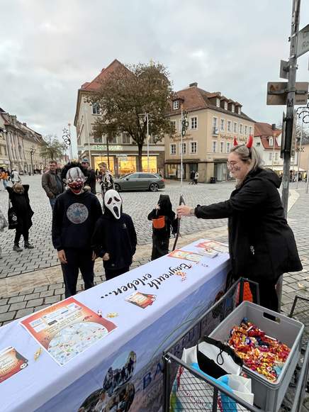 Eine Frau mit Teufelshörnern verteilt an einem Tisch im Freien auf einem Stadtplatz Süßigkeiten an Kinder in Halloween-Kostümen.