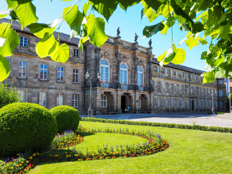 Ein historisches Steingebäude in der Welt der Wilhelmine mit einem von grünem Laub umrahmten Innenhof, der sich über einem runden Blumenbeet inmitten einer Rasenfläche erstreckt.
