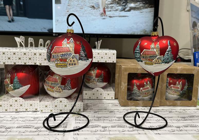 An schwarzen Ständern hängen zwei rote Weihnachtskugeln mit verschneiten Dorfmotiven. Dahinter stehen auf einem mit Notenblättern bedeckten Tisch Schachteln aus dem Bayreuther Shop, im Hintergrund ist ein Fernsehbildschirm zu sehen.