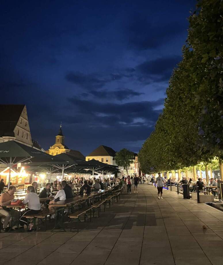 Menschen, die bei Einbruch der Dämmerung auf Tischen im Freien unter Sonnenschirmen bei einem Weinfest auf einem von Bäumen gesäumten Stadtplatz speisen.